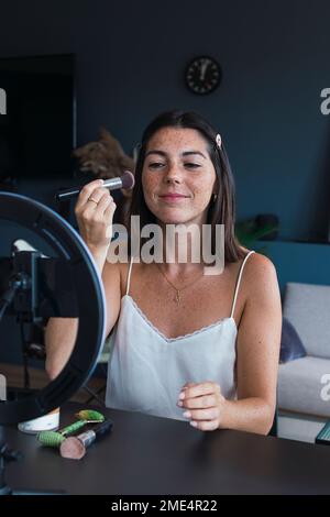 Jeune femme souriante appliquant le maquillage et le tutoriel de vlogging sur le smartphone à la maison Banque D'Images