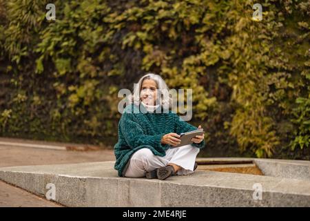 Femme âgée heureuse assise avec un PC tablette devant les plantes Banque D'Images