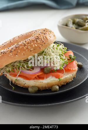 Saumon fumé frais et sain sur bagel de graines de sésame avec fromage à la crème, oignon rouge, pousses et câpres sur plaque texturée noire en format vertical. Banque D'Images