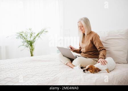 Femme mature utilisant un ordinateur portable et un chien de course sur le lit à la maison Banque D'Images