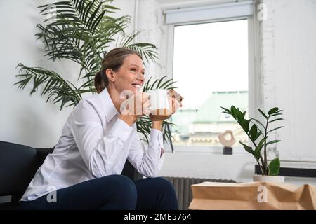 Bonne femme d'affaires mûre buvant du café assis sur un canapé dans le bureau Banque D'Images