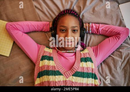 Vue de dessus portrait d'une adolescente noire qui écoute de la musique avec un casque tout en étant posée sur le lit et portant du rose Banque D'Images