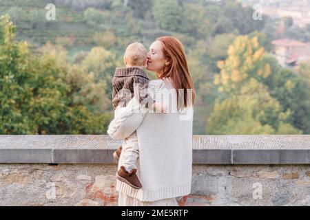 Une jeune femme aux cheveux rouges tient son tout-petit fils dans ses bras et l'embrasse tendinement. Ils se tiennent avec leur dos sur la toile de fond du vert hil Banque D'Images