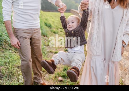 Maman et papa tiennent les bras du bébé et le roulent, il rit et balance ses jambes d'avant en arrière. Le garçon est vêtu d'un pull boutonné marron Banque D'Images