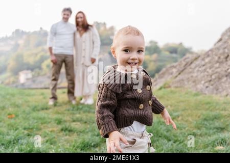 Un tout-petit garçon aux yeux bruns est en apparence souriant. Il est vêtu d'un pull tricoté brun à boutons, debout sur l'herbe verte avec ses parents qui m'embrasse Banque D'Images