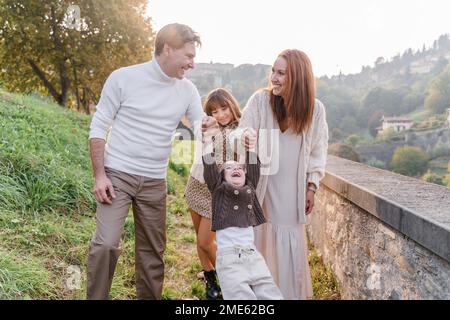 Maman et papa tiennent le tout-petit garçon par les mains et le roulent, il rit et balance ses jambes d'avant en arrière. Le garçon est vêtu d'un bouton tricoté marron Banque D'Images