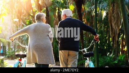 Nous continuons à être actifs. Vue arrière d'un couple âgé qui pousse ses vélos ensemble à l'extérieur dans un parc. Banque D'Images