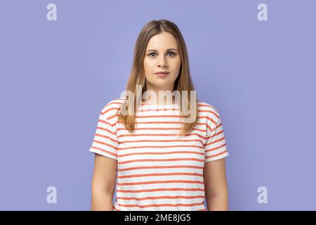 Portrait d'une femme blonde sérieuse et confiante portant un T-shirt rayé debout regardant l'appareil photo avec un look attentif, étant stricte et bossy. Studio d'intérieur isolé sur fond violet. Banque D'Images