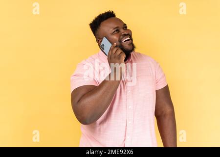 Portrait d'un homme joyeux portant une chemise rose parlant avec un ami sur un téléphone cellulaire, regardant loin et souriant joyeusement, ayant une conversation agréable. Studio d'intérieur isolé sur fond jaune. Banque D'Images