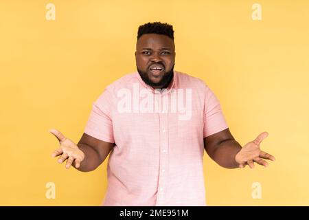Portrait d'un homme stressé malheureux portant une chemise rose debout bras levés et demandant, exprimant des émotions négatives, criant et frognant visage. Studio d'intérieur isolé sur fond jaune. Banque D'Images
