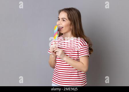 Portrait d'une petite fille portant un T-shirt rayé tenant des bonbons colorés, léchant, regardant loin avec l'expression childish satisfait. Prise de vue en studio isolée sur fond gris. Banque D'Images