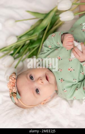 portrait d'une petite fille dans un serre-tête floral et d'un body vert dans un lit à la maison avec des tulipes. Fête des mères, 8 mars, printemps. Photo de haute qualité Banque D'Images