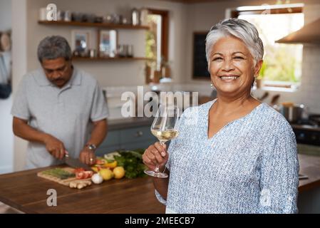 Dîners hubby ce soir. Portrait d'une femme mûre heureuse appréciant un verre de vin tandis que son mari prépare un repas en arrière-plan. Banque D'Images