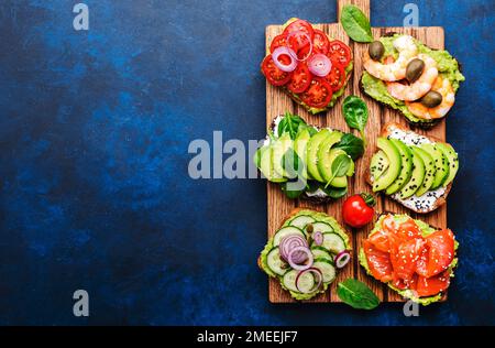 Set de sandwiches ou toasts à l'avocat : saumon, crevettes, tomates, concombres, fromage doux et épinards, noix de cajou et graines de sésame servis sur planche à découper, Banque D'Images