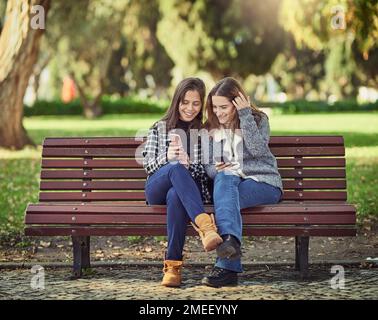 Jetez un coup d'œil à ce sujet. Prise de vue en longueur de deux jeunes femmes attirantes utilisant leur téléphone portable tout en étant assis dans le parc. Banque D'Images
