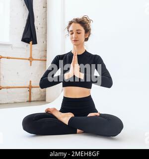 Jeune femme attrayante pratiquant le yoga, faisant la méditation avec les yeux fermés, assis en position de lotus avec namaste, s'exerçant seul dans le sport noir Banque D'Images