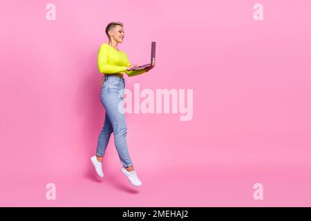 Photo pleine grandeur de la jeune femme attrayante saut à pied ordinateur portable ouvrier habillé élégant jaune vêtements isolés sur fond rose couleur Banque D'Images