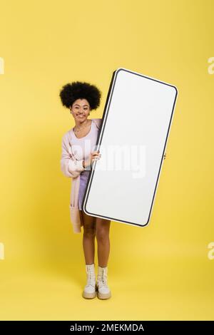 pleine longueur de gaie afro-américaine femme en cuir bottes tenant un énorme modèle de smartphone sur fond jaune Banque D'Images