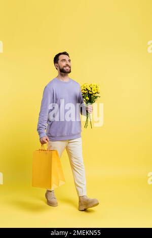 pleine longueur d'homme heureux et élégant marchant avec des sacs de shopping et des fleurs sur fond jaune Banque D'Images