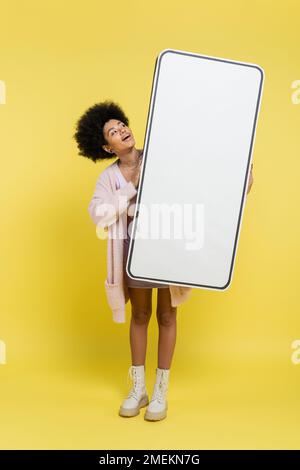 pleine longueur de femme afro-américaine stupéfait tenant un énorme modèle de carton de smartphone sur fond jaune Banque D'Images