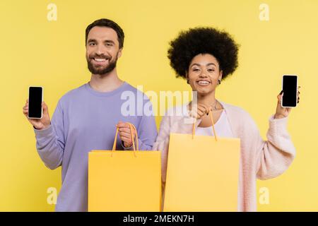 joyeux couple multiethnique avec des sacs de shopping montrant des téléphones mobiles avec écran vierge isolé sur jaune Banque D'Images