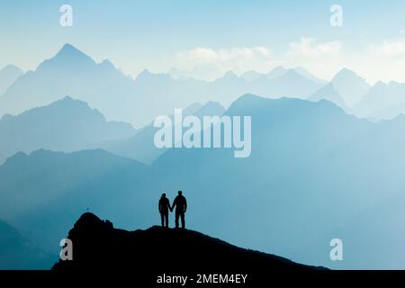Silhouette couple d'homme et de femme atteignant le sommet de la montagne jouissant de la liberté et regardant vers les silhouettes de montagne bleues et le lever du soleil. Alpes, Allgaeu Banque D'Images