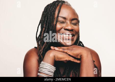 Femme heureuse avec des dreadlocks souriant avec ses yeux fermés sur un fond blanc. Joyeuse femme mûre portant un maquillage léger et des bijoux. Joyeux Banque D'Images