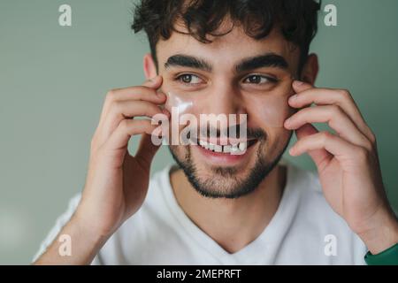 Portrait de l'homme souriant regardant loin appliquer de la crème hydratante sur son visage isolé sur fond de mur vert. Concept de routine de soin de la peau. Banque D'Images