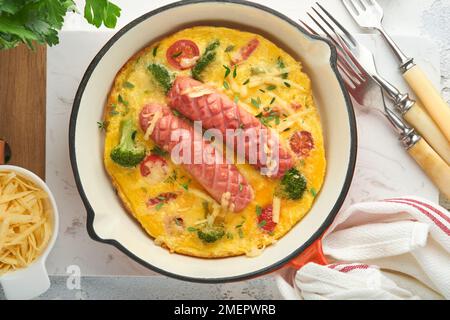 Omelette ou frittata avec deux saucisses, fromage fondu, tomates cerises, brocoli et persil sur poêle rouge ou poêle en fer sur fond blanc en béton Banque D'Images