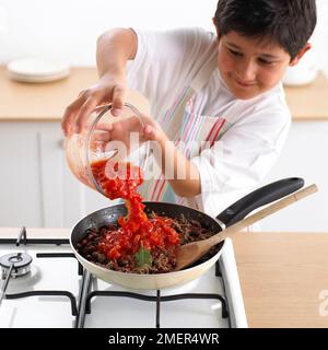 Garçon ajoutant des tomates hachées au bœuf haché frit, 9 ans Banque D'Images