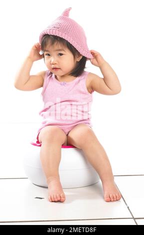 Fille portant un gilet rose assis sur un pot blanc, portant un chapeau de laine rose, 20 mois Banque D'Images