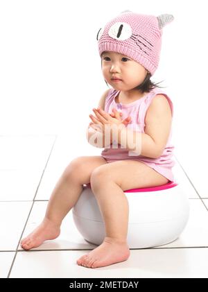 Fille portant un gilet rose et un chapeau en laine rose, assise sur un pot blanc, 20 mois Banque D'Images