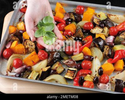 Ajouter des olives et des feuilles de basilic frais aux légumes rôtis (courgettes, aubergines, oignons, poivrons rouges, tomates cerises, courge musquée, ail) Banque D'Images