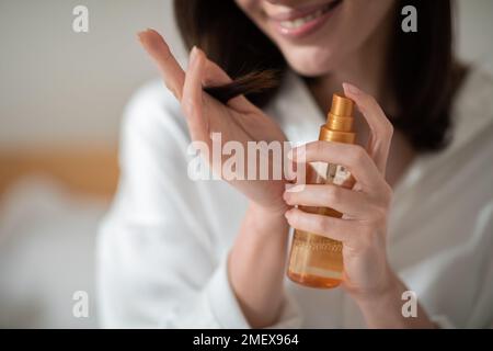 Gaie jeune femme européenne sur lit blanc applique un spray sur les cheveux dans la chambre à coucher intérieur, gros plan, court Banque D'Images