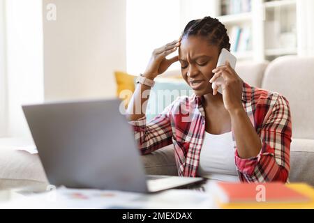 Une femme africaine en difficulté s'assoit sur le lieu de travail, en utilisant un ordinateur, un téléphone Banque D'Images