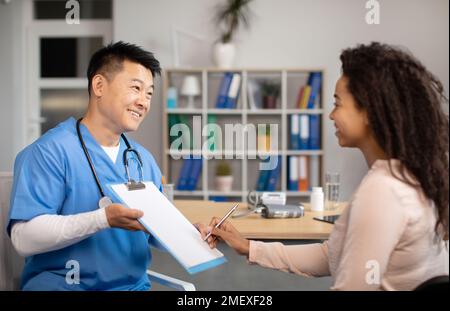 Heureux homme coréen d'âge moyen médecin donnant le contrat de signature à la patiente noire millénaire en clinique Banque D'Images