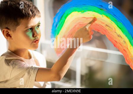 Petit garçon sérieux dessine l'arc-en-ciel sur la fenêtre avec la main, rêve, pensez à l'intérieur de la chambre, lumière du soleil, gros plan Banque D'Images