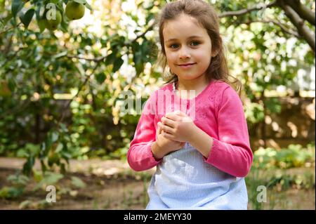 Un adorable enfant caucasien, une petite fille de 5 ans sourit en regardant la caméra, tenant une poire mûre fraîche dans ses mains, debout seule dans un verger. Harv Banque D'Images