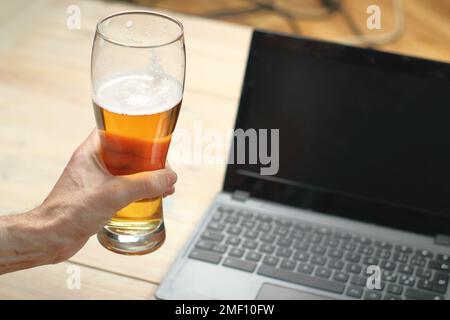 personne tenant une tasse à bière près d'un ordinateur portable sur une table en bois Banque D'Images