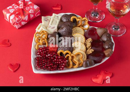 Saint-Valentin assiette de bonbons coeur en forme de coeur avec chocolat, bonbons en gelée, crackers, cookies, noix, grenade et deux verres de vin rose sur la surface rouge Banque D'Images