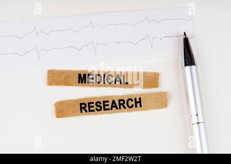 Concept médical. Sur le cardiogramme se trouve un stylo et un papier déchiré avec le texte - RECHERCHE MÉDICALE Banque D'Images