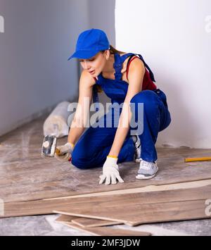 Femme concentrée dans la construction bleue combinaisons pose de revêtement de sol stratifié, frappe avec un marteau souple sur la pièce avec verrou pour la fixation Banque D'Images