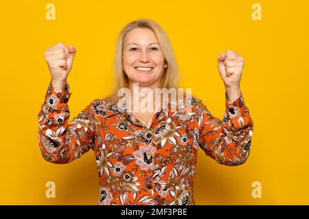 Bonne femme caucasienne réussie dans une robe à motifs avec des mains en relief souriant et célébrant le succès sur fond jaune. Banque D'Images