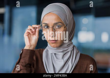Gros plan. Portrait de la jeune belle femme arabe dans le hijab. Il regarde avec confiance dans l'appareil photo, soutient ses lunettes avec sa main. Banque D'Images