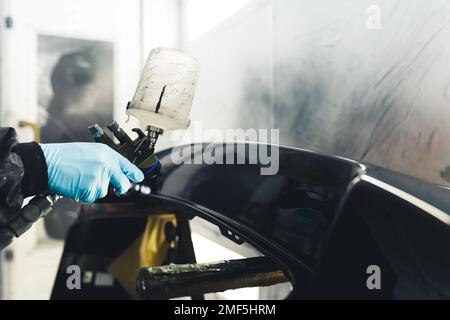 Gros plan de la personne portant des gants bleus peignant une partie de la voiture noire à l'aide d'un aérographe. Peinture automobile. Prise de vue horizontale en intérieur. Photo de haute qualité Banque D'Images