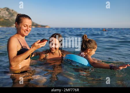 Une jeune mère heureuse et ses deux filles gaies dans des maillots de bain lumineux avec un petit cercle gonflable bleu prendre des photos pour la mémoire, dans le summe azur Banque D'Images