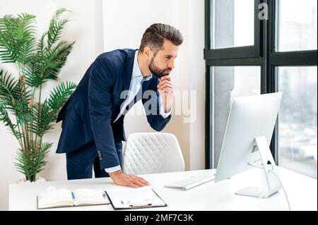 Homme d'affaires caucasien, pdg de la société, directeur financier, se tient près du bureau dans l'élégant costume, travaillant dans l'ordinateur, concentré conduit la correspondance de travail en ligne Banque D'Images