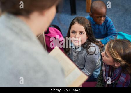 Ils écoutent toujours très attentivement lors de son histoire. un groupe d'enfants de l'école élémentaire qui écoutent leur professeur lisent une histoire. Banque D'Images