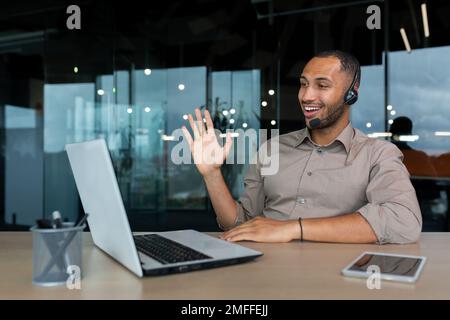 Formation en ligne. Un jeune étudiant afro-américain est assis dans un bureau, à un bureau, devant un ordinateur portable, dans un casque. Apprend par un appel vidéo, dit bonjour, sourit. Banque D'Images