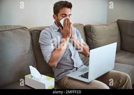 Même si je suis malade, je dois continuer à travailler. un jeune homme qui regarde mal à l'aise éternuant dans un tissu tout en essayant de travailler sur son ordinateur portable à la maison. Banque D'Images